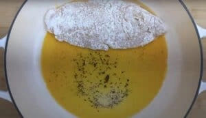 Floured chicken breast in oil with spices in a white pan, ready for cooking.